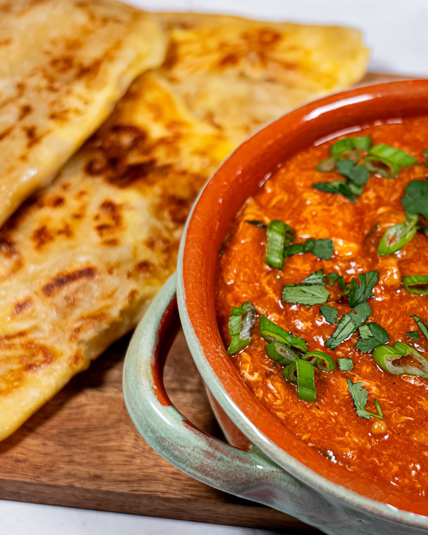 A bowl of sweet and spicy chili chicken garnished with fresh cilantro and green onions, served with crispy Roti Prata on a wooden board.