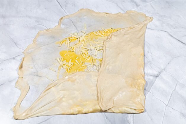 Thinly stretched dough with beaten egg and shredded cheese, partially folded.