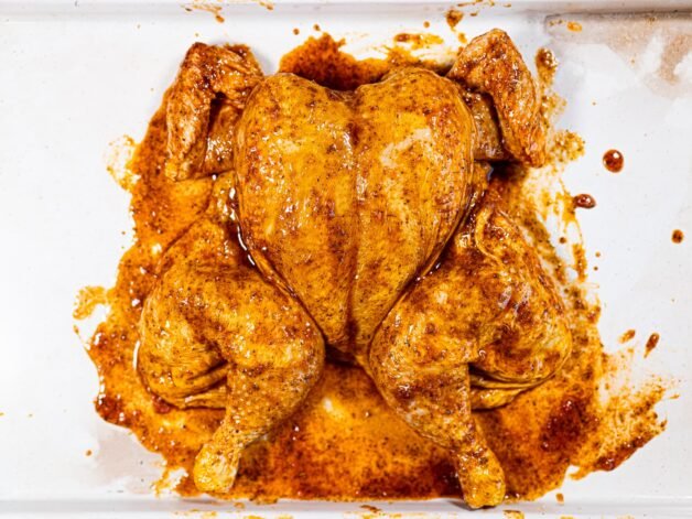 Spatchcocked chicken fully coated in a smoky, herb-infused marinade, resting in a white roasting pan before cooking.