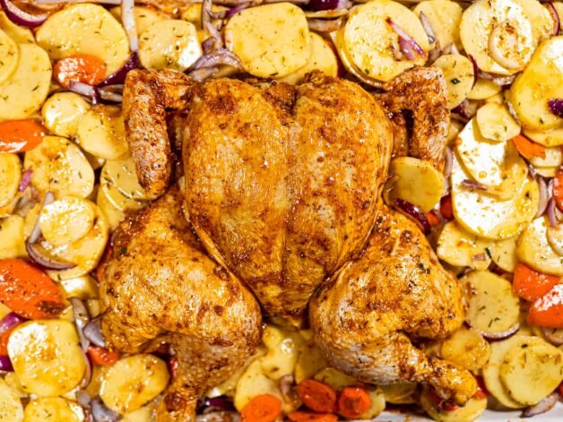 Spatchcocked chicken coated in marinade, placed over a bed of seasoned potatoes, carrots, and red onion in a roasting pan, ready for the oven.
