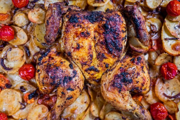 Close-up top view of spatchcock roasted chicken with crispy golden skin, surrounded by roasted potatoes, cherry tomatoes, and caramelized onions.