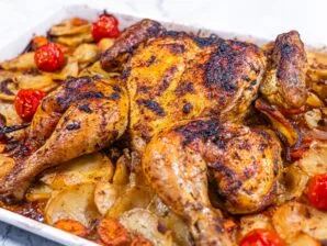 Golden-brown spatchcock roasted chicken with crispy skin, resting on a bed of roasted potatoes, carrots, red onion, and cherry tomatoes in a white baking dish.