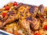 Golden-brown spatchcock roasted chicken with crispy skin, resting on a bed of roasted potatoes, carrots, red onion, and cherry tomatoes in a white baking dish.