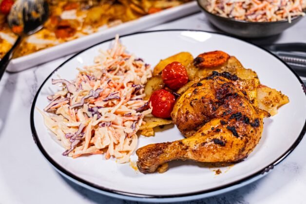 A plate of crispy spatchcock roasted chicken with golden roasted potatoes, cherry tomatoes, and a side of creamy coleslaw, served on a white dish.