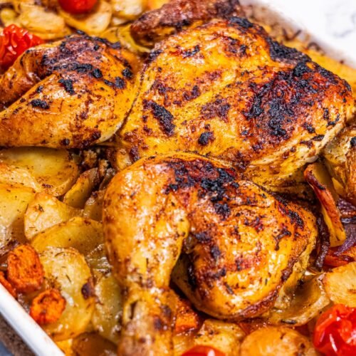 Golden-brown spatchcock roasted chicken with crispy skin, served over a bed of roasted potatoes, carrots, and cherry tomatoes in a white baking dish.