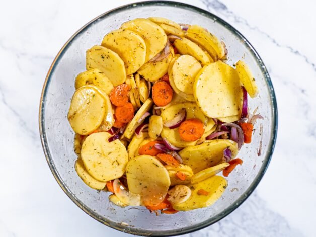 Sliced potatoes, carrots, and red onion mixed with seasoning and olive oil in a glass bowl, ready for roasting.