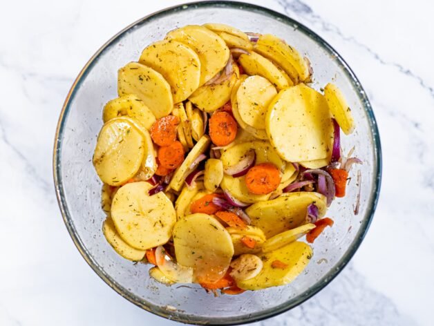 Sliced potatoes, carrots, and red onion mixed with seasoning and olive oil in a glass bowl, ready for roasting.