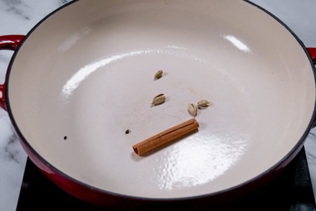 Whole cinnamon stick and green cardamom pods toasting in a dry pan to enhance their aroma and depth of flavor.